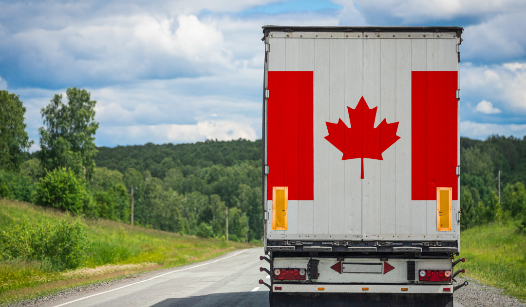 A delivery truck with the Canadian flag on the back drives down a road, surrounded by green trees.