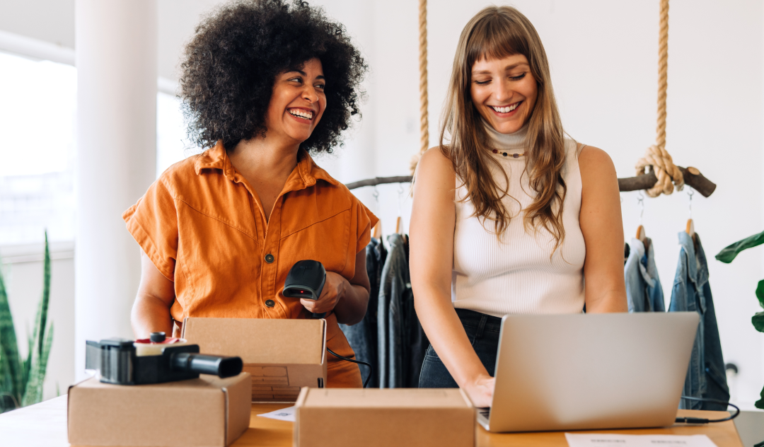 Two girls stand beside each other smiling as they work together to scan and package shipments.